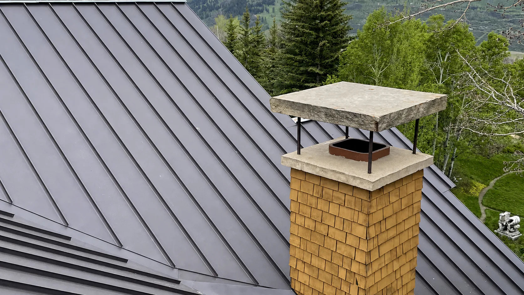 Completed dark standing seam metal roof beside a cedar-shingle chimney in a mountain setting